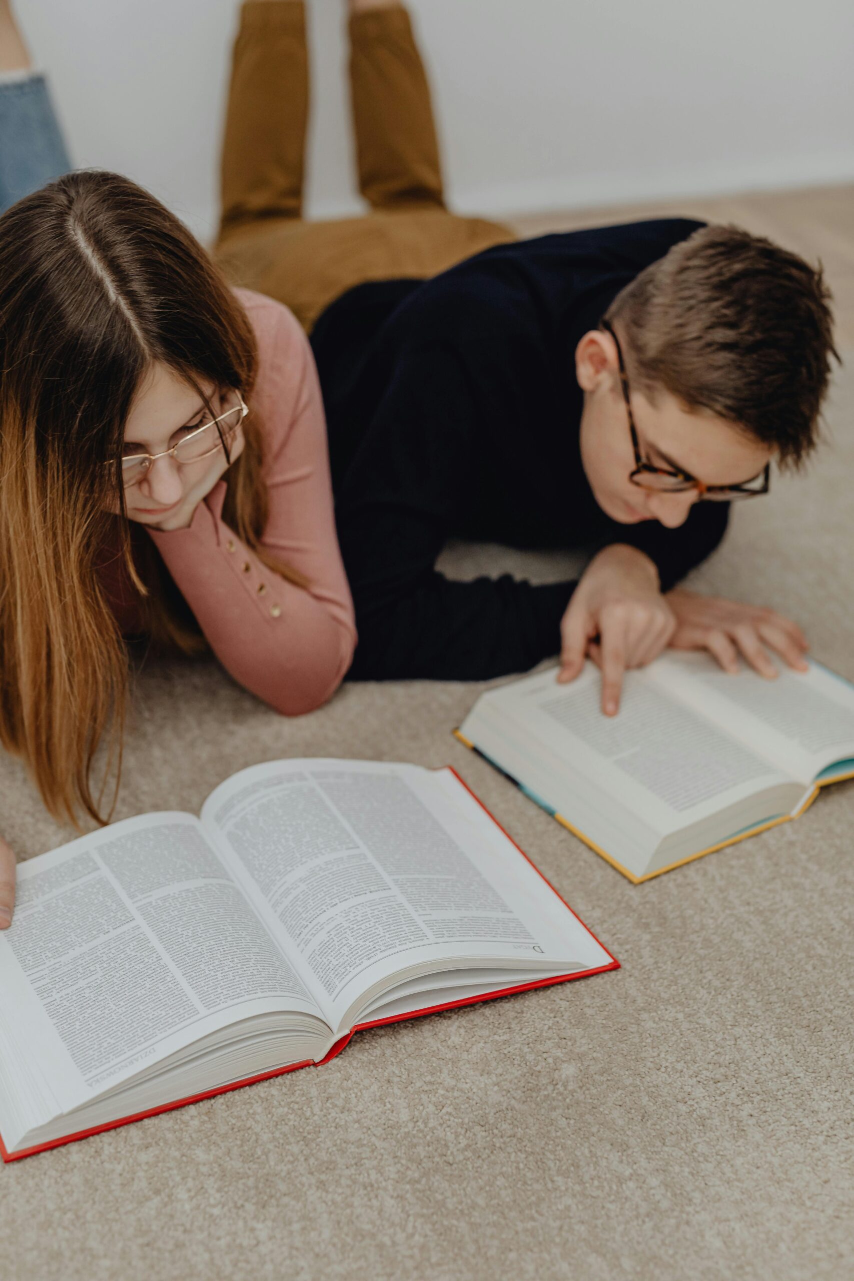 Two people laying on their fronts on the floor while each reading a book.