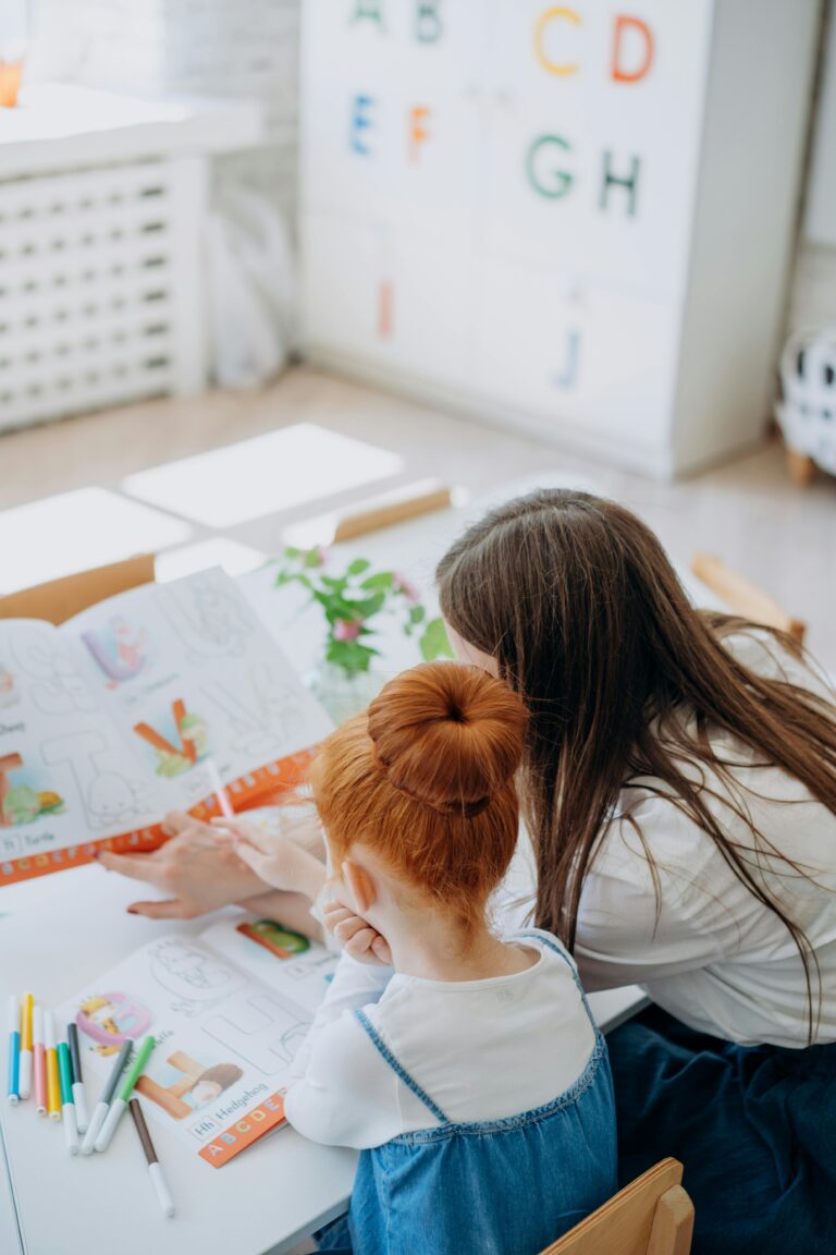 From behind, a mom and daughter are sitting next to each other with their heads leaning towards each other while reading a book.