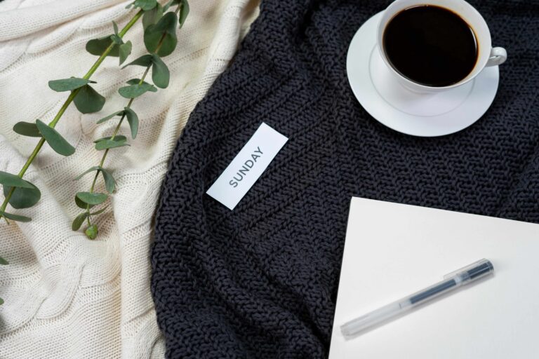 Cozy textiles in the backgound of the flatlay. Laying on top is eucalyptus, a notepad, a cup of coffee, and a slip of paper with "Sunday" on it