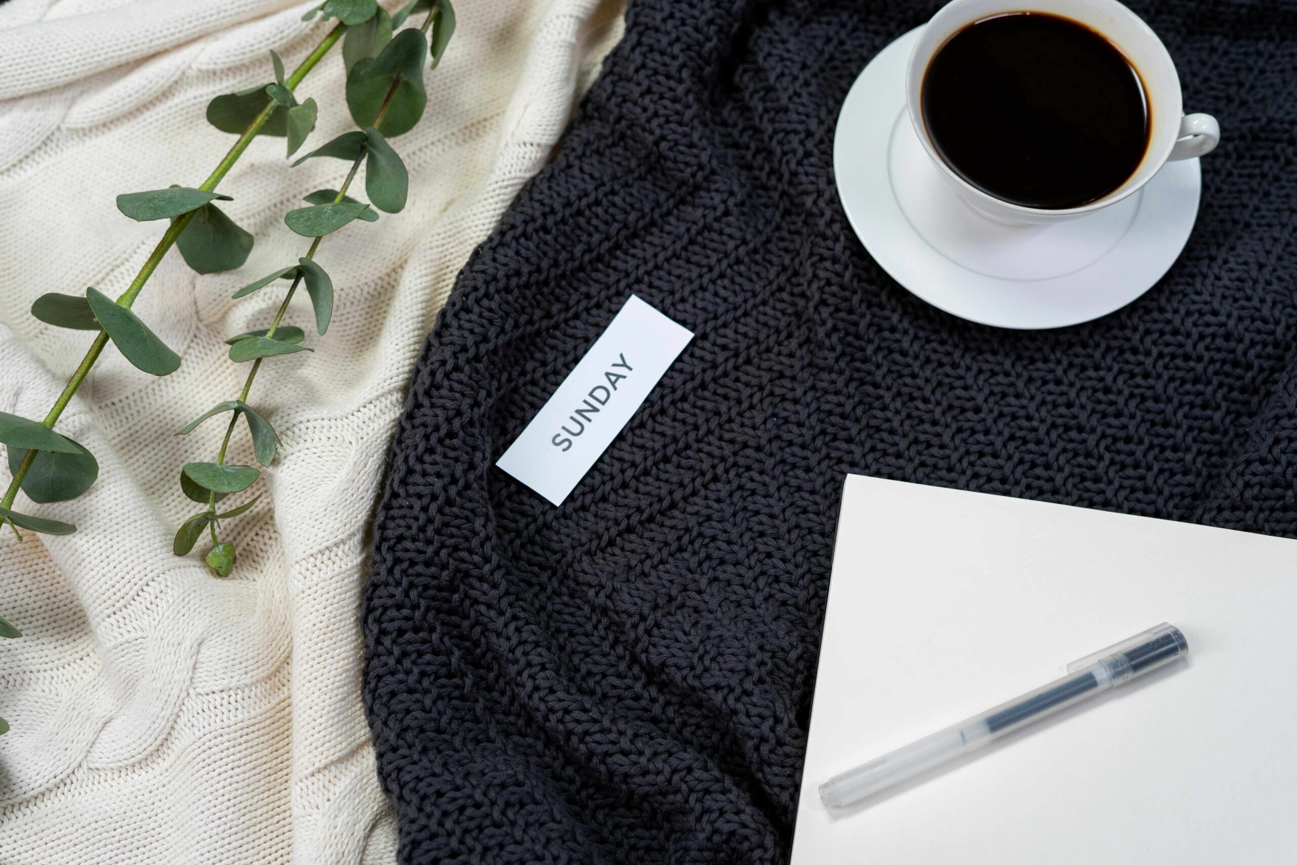 Cozy textiles in the backgound of the flatlay. Laying on top is eucalyptus, a notepad, a cup of coffee, and a slip of paper with "Sunday" on it